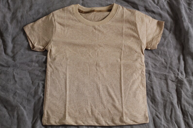 May include: A short-sleeved, tan-colored t-shirt. The shirt has a crew neck and is made of a soft, textured fabric. The t-shirt is laying flat on a gray, wrinkled surface.