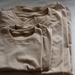 May include: Three folded, tan-colored t-shirts of varying sizes are stacked on top of each other. The shirts are made of a soft material and have a classic crew neck design. The shirts are neatly arranged on a light gray surface.