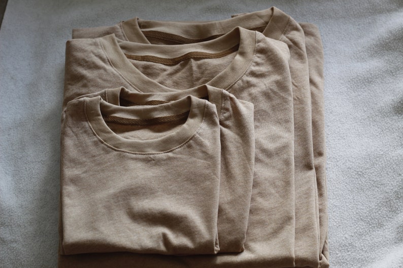 May include: Three folded, tan-colored t-shirts of varying sizes are stacked on top of each other. The shirts are made of a soft material and have a classic crew neck design. The shirts are neatly arranged on a light gray surface.