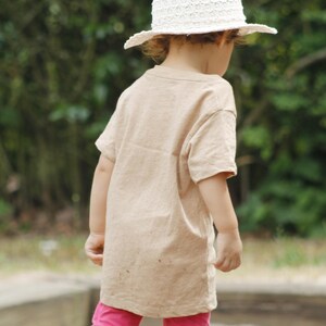 May include: A child wearing a beige t-shirt, pink leggings, and a white sun hat. The hat has a wide brim and a textured design. The child is standing outdoors with a blurred green background.