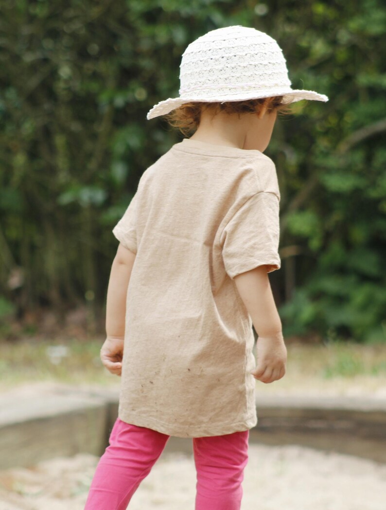 May include: A child wearing a beige t-shirt, pink leggings, and a white sun hat. The hat has a wide brim and a textured design. The child is standing outdoors with a blurred green background.
