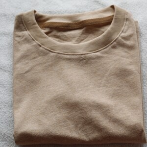 May include: Folded tan t-shirt with a crew neck. The shirt is made of a soft material and is neatly folded. The neutral color makes it a versatile wardrobe staple. The shirt is ready to wear.