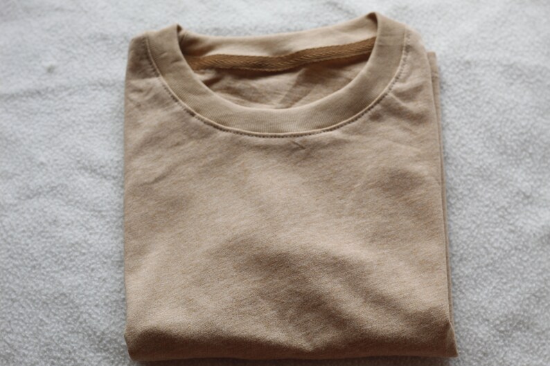 May include: Folded tan t-shirt with a crew neck. The shirt is made of a soft material and is neatly folded. The neutral color makes it a versatile wardrobe staple. The shirt is ready to wear.