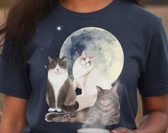 May include: A navy blue t-shirt featuring a watercolor illustration of three cats in front of a full moon. The person in the image is holding a beige mug. The background is a field with a full moon in the sky.