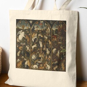 Könnte beinhalten: Eine cremefarbene Tragetasche mit einem detaillierten, mehrteiligen Kunstwerk, das zahlreiche Figuren und Tiere zeigt. Die Tasche hat lange Henkel und steht auf einer Holzoberfläche, mit einem Korb und einer Pflanze im Hintergrund.
