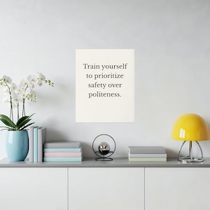 May include: A white wall art print with the text "Train yourself to prioritize safety over politeness." A light blue vase with white flowers sits to the left. A yellow lamp and decorative items are on a white cabinet.
