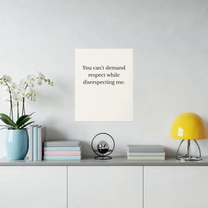 May include: A framed print with the text "You can't demand respect while disrespecting me." The print is cream-colored and mounted on a white wall. A yellow lamp, books, and a decorative object are on a gray shelf below.
