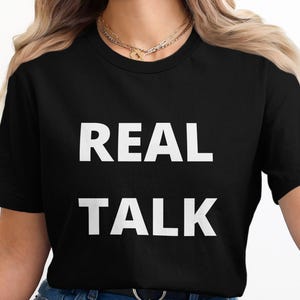 May include: Black t-shirt with the words "REAL TALK" printed in large, white capital letters. The shirt is a classic crew neck style.