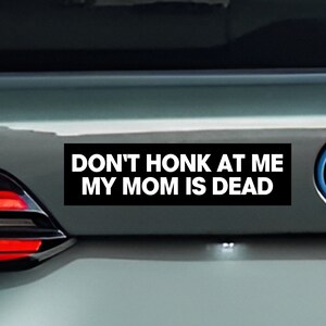 Op de afbeelding: Zwart-witte bumper sticker met de tekst "DON'T HONK AT ME MY MOM IS DEAD" op een zilveren auto.