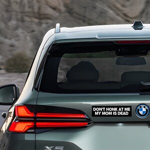 Op de afbeelding: Een zilveren auto met een zwarte bumper sticker waarop staat "Niet toeteren, mijn moeder is dood".