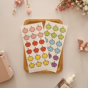 Könnte beinhalten: Ein Bogen mit bunten Apfel-Stickern mit verschiedenen Gesichtsausdrücken. Die Sticker sind in Reihen zu vieren angeordnet, wobei jede Reihe eine andere Farbe hat. Die Farben umfassen Rosa, Rot, Orange, Gelb, Grün, Blau und Lila. Die Sticker befinden sich auf einem Holzgrund.