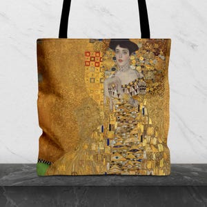 May include: A tote bag featuring a reproduction of Gustav Klimt's Portrait of Adele Bloch-Bauer I. The design displays a figure in a gold-toned dress with geometric patterns, intricate textures, and dark hair, set against a shimmering gold background.