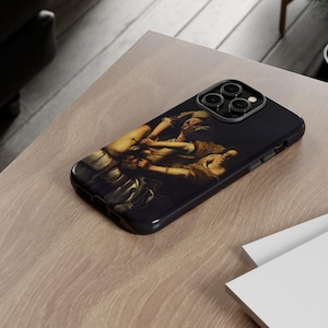 May include: Black iPhone case featuring a classical painting of figures in shades of brown, beige, and white. The case has a glossy finish and a camera cutout. The phone case is resting on a light brown wooden surface.