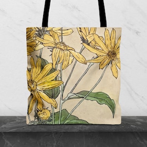 Peut inclure: Un sac fourre-tout orné d'une illustration botanique de fleurs jaunes avec des feuilles vertes sur un fond beige clair. Le motif présente des contours noirs et des tiges fines, avec deux anses en tissu noir partant du haut du sac.
