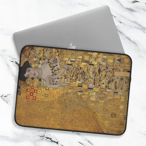 May include: A rectangular laptop sleeve featuring a printed reproduction of Gustav Klimt's painting Adele Bloch-Bauer I. The design displays a portrait of a person surrounded by intricate gold, yellow, and geometric patterns with a black border.