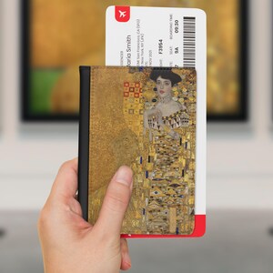 May include: A passport holder featuring a gold-toned reproduction of Gustav Klimt's Portrait of Adele Bloch-Bauer I, with a black spine. An airline boarding pass for Maria Smith, flight F3954, seat 9A, is tucked inside the rectangular cover.