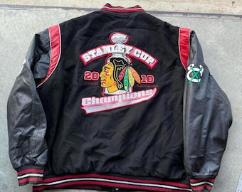 Może przedstawiać: Czarna satynowa kurtka z czerwonymi i białymi wykończeniami, z naszywką Stanley Cup Champions. Naszywka zawiera logo drużyny, lata 2010 i 2010 oraz słowo "Champions". Rękawy są czarne z fakturą przypominającą skórę.