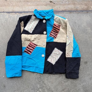 May include: A patchwork jacket in a variety of colors including black, blue, tan, and pink. The jacket has a collared neckline and is fastened with white buttons. Decorative patches with embroidered details are sewn onto the jacket.