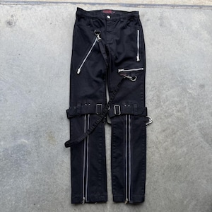 May include: Black pants with silver zippers running vertically along the front of each leg. The pants have multiple silver metal accents, including D-rings and a chain. Adjustable straps and buckles are also visible.