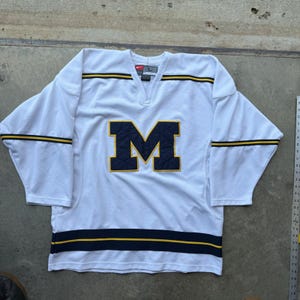 May include: A white long-sleeve hockey jersey featuring a large navy blue letter M with a quilted texture on the chest. The jersey has navy blue and yellow horizontal stripes on the sleeves and hem, a V-neck collar, and a Nike logo on the inner label.