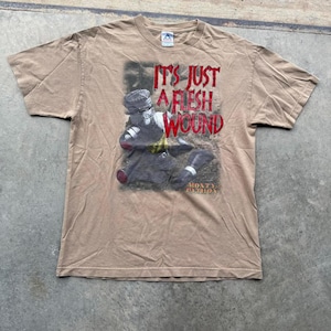 Peut inclure: T-shirt beige à manches courtes avec un motif de chevalier et le texte rouge "IT'S JUST A FLESH WOUND". Le t-shirt comprend également le texte "MONTY PYTHON" en plus petit en dessous de l'image du chevalier.