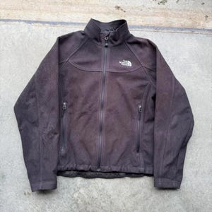 May include: A dark brown The North Face full-zip jacket. The jacket has a high collar, two side pockets with zippers, and a small logo on the chest. The jacket is made of a soft, textured material.