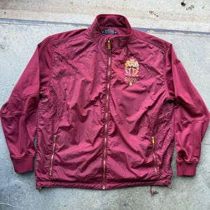 May include: A burgundy zip-up jacket featuring a high collar, long sleeves with ribbed cuffs, and two front pockets with gold-tone zippers. The chest displays an embroidered crest logo with the text COOGI, and tonal intricate patterns.