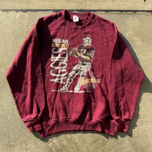 May include: A maroon long-sleeve crewneck sweatshirt featuring a graphic of a football player in a helmet and uniform. The text reads Texas A&M Aggies and Champions Conference. The design includes a football and player in a dynamic pose.