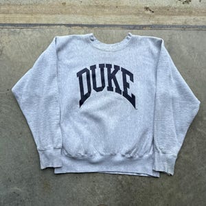 May include: A heather gray long-sleeve crewneck sweatshirt featuring the word DUKE printed in large, dark navy blue capital letters across the chest in an arched font. The garment has ribbed cuffs and a ribbed hemline at the bottom edge.