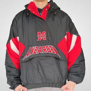 May include: Black and red hooded anorak jacket with the word "NEBRASKA" in white lettering. The jacket features a red lining and a red "M" logo. The jacket has a drawstring hood and a front pocket.