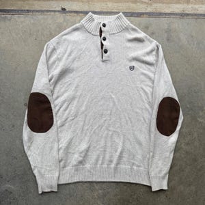 Op de afbeelding: Een lichtbeige trui met een opstaande kraag, een knoopsluiting met drie donkerbruine knopen, lange mouwen met donkerbruine elleboogstukken en een klein geborduurd embleemlogo op de linkerborst. De boorden en de zoom zijn afgewerkt met ribbelbreisel.