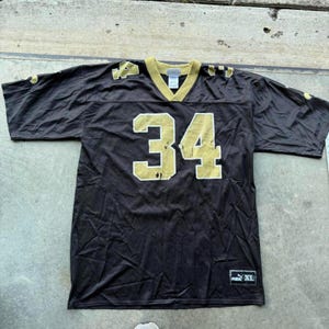 Peut inclure: Un maillot de football noir et or avec le numéro 34. Le maillot a un col en V et des manches courtes. Le logo Puma et l'étiquette de taille XL sont visibles en bas à droite.