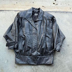 May include: Black leather jacket with a button-up front, two pockets, and a wide, banded bottom. The jacket has a collar and short sleeves. The brand name "A. COMANI" is visible on the label. The jacket is made of a shiny, possibly vintage leather.