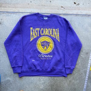 Può includere: Una felpa viola a maniche lunghe con girocollo caratterizzata da una scritta serigrafata in giallo che recita East Carolina, Established 1907, e Pirates, Greenville, NC. Un sigillo universitario circolare è centrato sul petto con un'etichetta Lee.