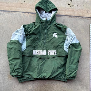 Puede incluir: Chaqueta anorak verde y azul claro con capucha. La chaqueta presenta las palabras "MICHIGAN STATE" en letras blancas en la parte delantera, junto con un logotipo de casco espartano. La chaqueta tiene un cierre de media cremallera.