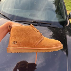 May include: A tan suede ankle boot with a lace-up closure and a flat rubber sole. The boot features a pull tab at the heel and the UGG logo embossed on the outer side. The design includes tonal stitching and a rounded toe, showcasing a simple construction.