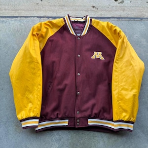 May include: A maroon and gold varsity jacket with a button-up front. The sleeves are gold, and the body is maroon. The jacket has a white, gold, and maroon striped collar and cuffs. A gold "M" logo is on the chest.