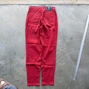 Puede incluir: Vaqueros rojos de mezclilla con un diseño clásico de cinco bolsillos. Los vaqueros tienen una pequeña etiqueta negra con texto blanco en la cintura trasera. Los vaqueros están colocados sobre una superficie de hormigón.