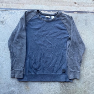 May include: A long-sleeve crewneck sweatshirt featuring a blue center panel and contrasting dark gray raglan sleeves. The garment includes a ribbed neckline, cuffs, and hem, with a small dark rectangular brand patch located on the lower right side.