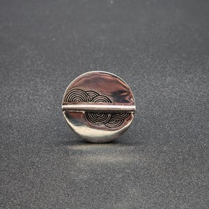 May include: A silver-toned, circular pin with a horizontal line bisecting the circle. The top half features a series of concentric, curved lines. The pin has a shiny, reflective surface and a simple, modern design.
