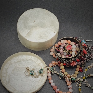 May include: Two white mother-of-pearl trinket dishes with a variety of colourful beaded bracelets and necklaces, including a silver turtle charm bracelet.