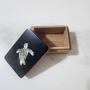 Può includere: Piccola scatola di legno con coperchio incernierato. Il coperchio nero presenta un motivo decorativo a tartaruga intarsiato con pezzi di conchiglia iridescenti. La scatola è aperta, rivelando l'interno in legno naturale.