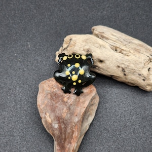 Puede incluir: Figura de rana de cerámica negra con manchas amarillas. La rana está sentada en una pequeña roca.