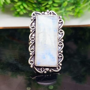 May include: A rectangular silver ring featuring a large, light-colored, rectangular moonstone. The moonstone has subtle blue and white hues. The ring's silver setting has an ornate, scrolling design around the stone, adding a vintage aesthetic.