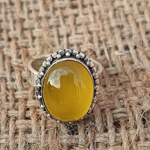 Könnte beinhalten: Ein silberner Ring mit einem gelben ovalen Edelstein, der in einer Perlenfassung sitzt. Der Ring liegt auf einem gewebten Stoffhintergrund.