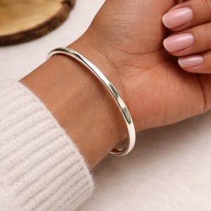 Puede incluir: Un brazalete de plata pulida usado en la muñeca. El brazalete tiene un diseño simple y redondeado. El tono de piel es medio y las uñas están pintadas de color rosa claro. La persona lleva un suéter blanco.