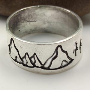 Op de afbeelding: Een zilveren ring met een bergketen- en boomontwerp. De ring heeft een brede band met een gedetailleerd berglandschap en twee bomen die rond de band zijn geëtst. De tekst "DevJewellrs" is ook zichtbaar.