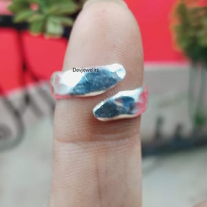 May include: A silver-colored ring with a hammered texture. The ring wraps around the finger with an open design. The text "Devjewellrs" is visible on the finger. This ring is a piece of jewelry.