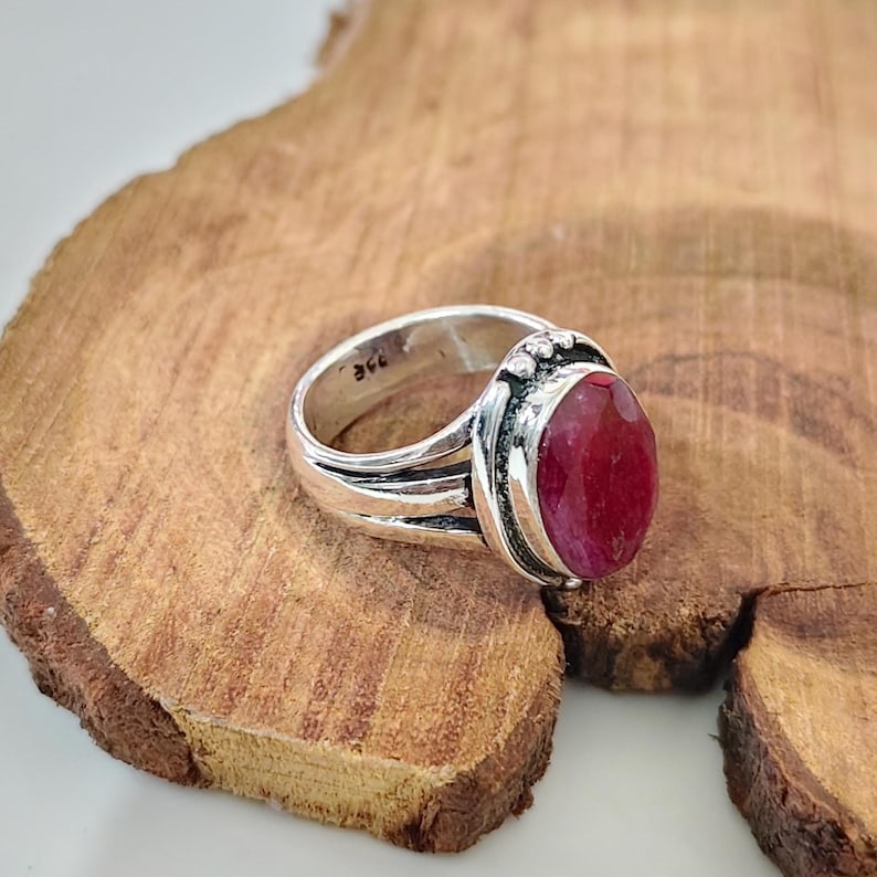 May include: A silver ring featuring a large, oval-shaped, faceted deep red gemstone set in a polished metal band. The band has a split-shank design and includes a small hallmark stamp visible on the inner surface of the silver metal ring.