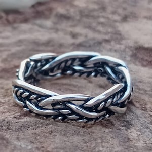 Puede incluir: Un anillo plateado con un diseño trenzado. El anillo tiene un aspecto texturizado, con hebras entrelazadas que crean un patrón detallado. El anillo está sobre un fondo neutro y texturizado.